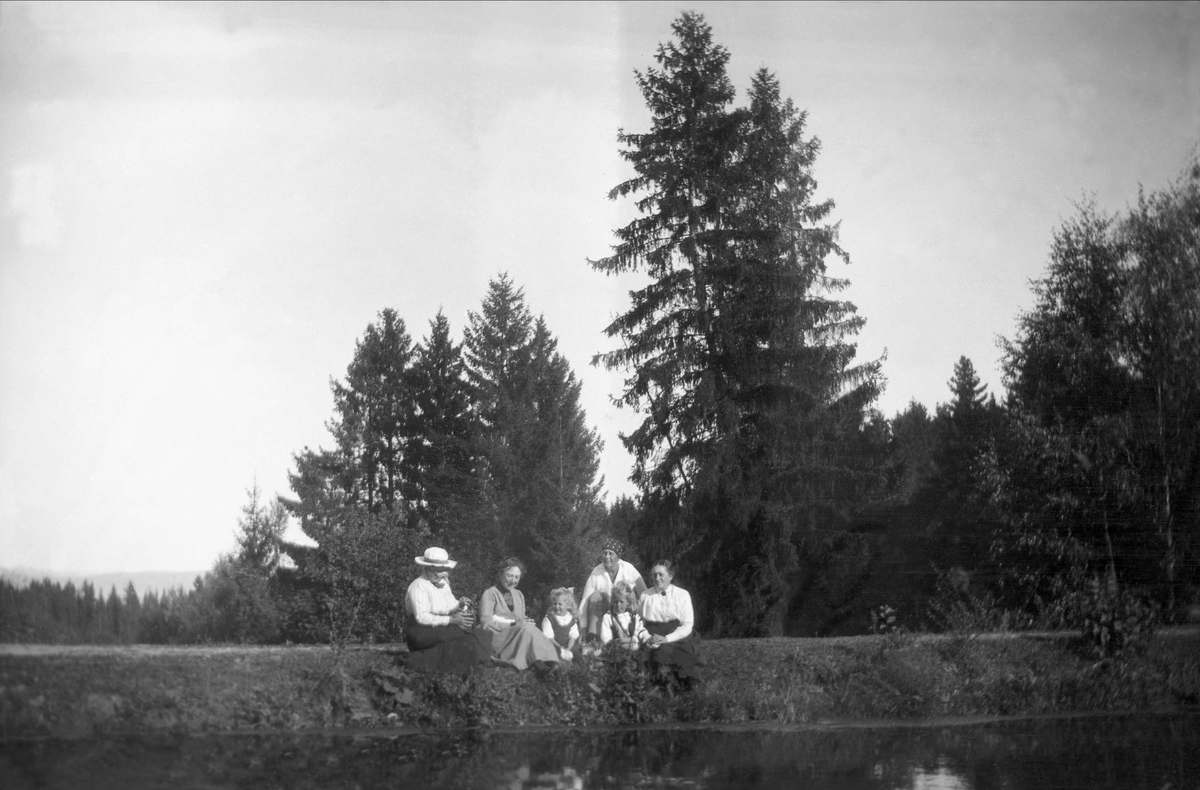 Fire kvinner og to barn sitter å poserer nede ved vannet i en skog. Robsahm og Lund.