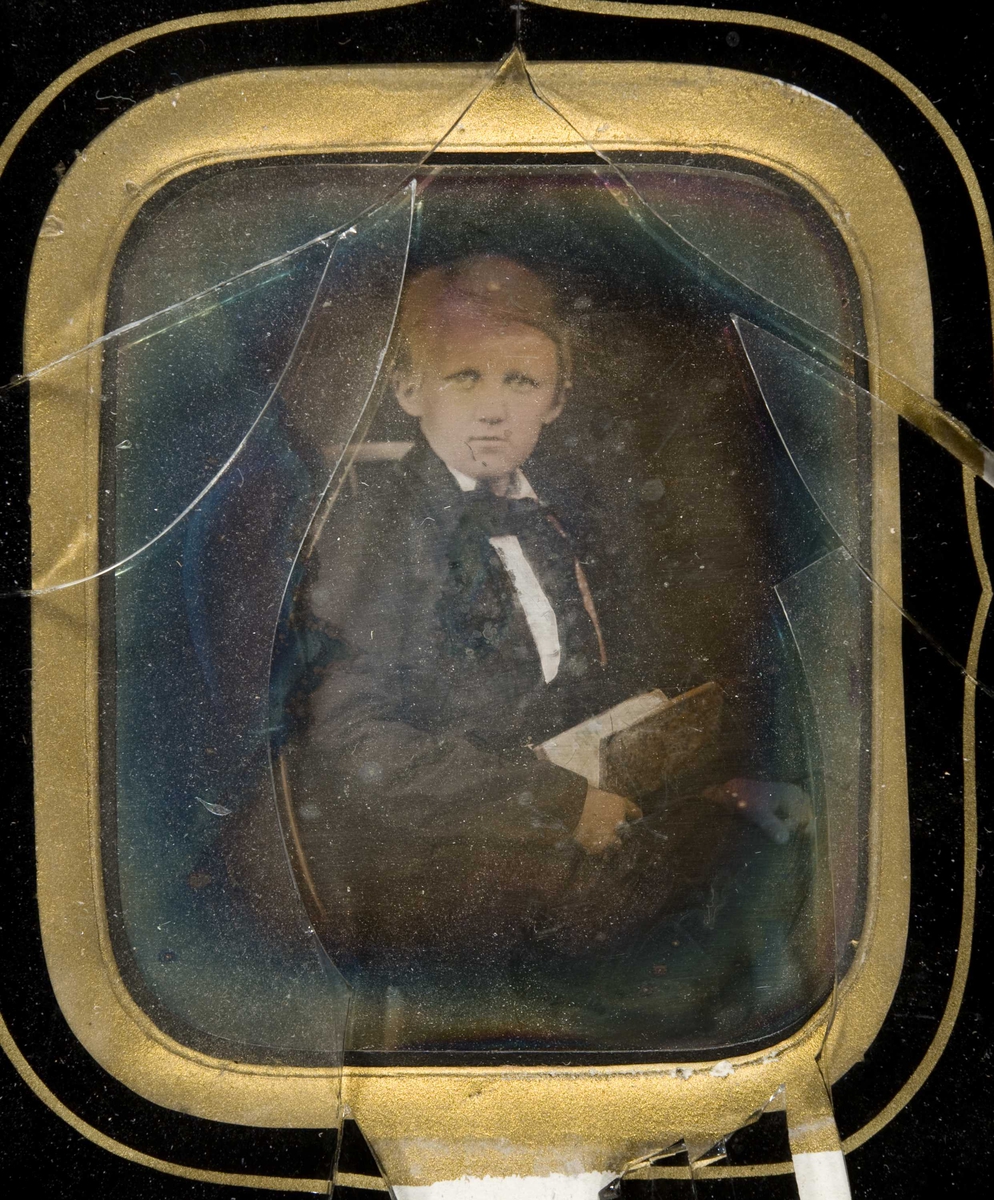 Daguerreotypi. Halvfigur portrett av ukjent gutt. Han sitter med en bok i fanget. Knust glass