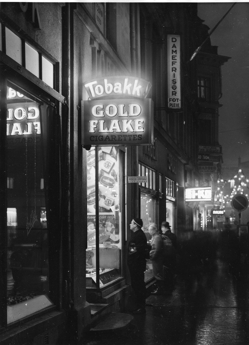 Gatebilde. Tre menn ser på vindusutstillinger, blant annet til en tobakksbutikk. Neonskilt med reklame for Gold Flake sigaretter.