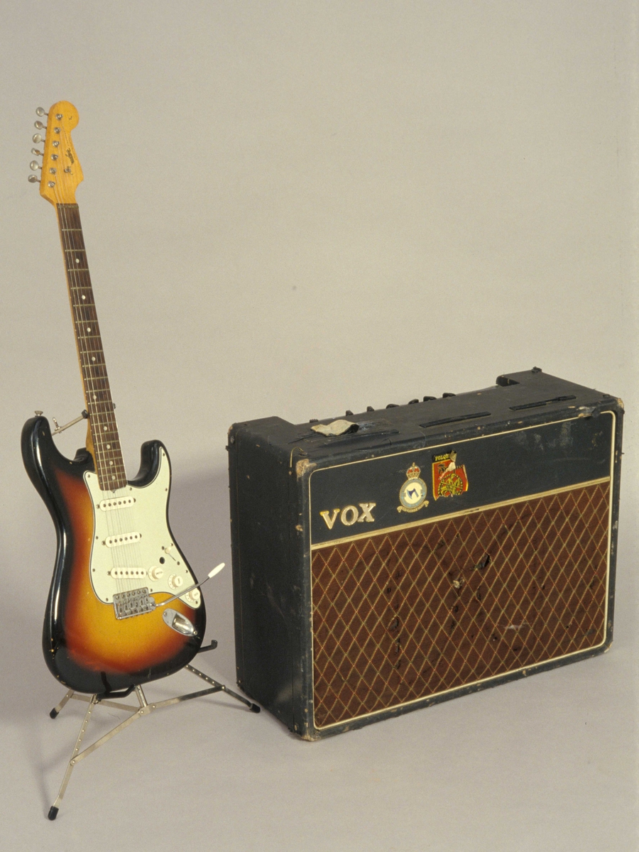 Elektrisk gitar, Fender Stratocaster, stående i gitarstativ, og en VOX AC30 forsterker.