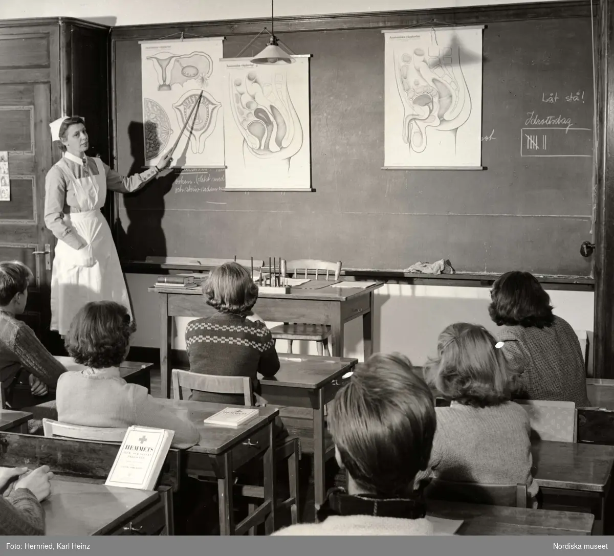 Sexualundervisning i skola. En kvinna i sköterskeuniform visar anatomiska planscher på svarta tavlan. Eleverna sitter vid sina skolbänkar.