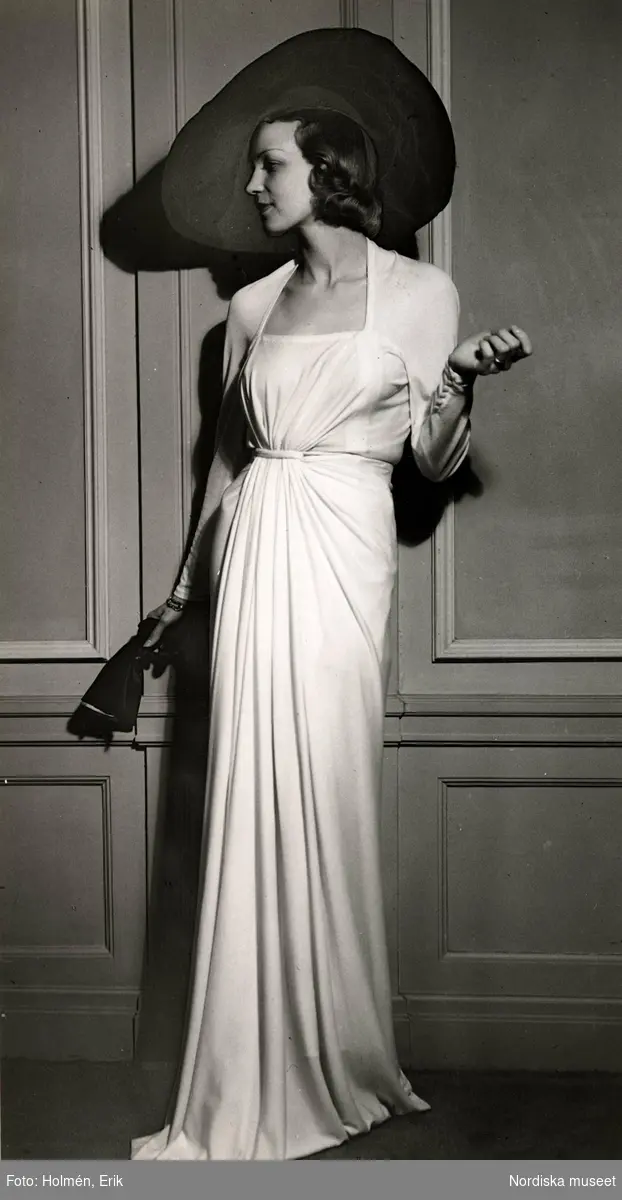 Dammode, kvinnlig modell poserar iklädd draperad aftonklänning och vidbrättad hatt. I handen håller hon ett par handskar. Nordiska Kompaniet 1938.