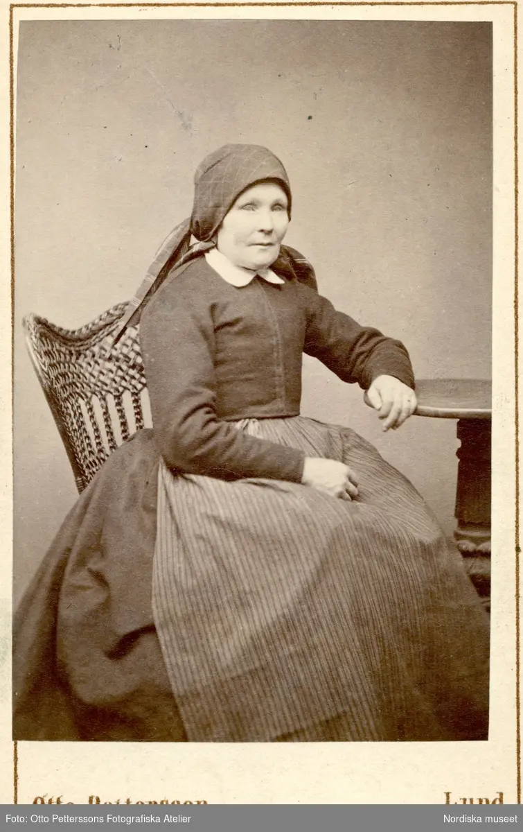 Porträtt av sittande kvinna iklädd sjalett och randigt förkläde. Hon vilar armen på ett pelarbord. Andrarum, Albo hd. Gåva av fotograf Otto Petterson, Lund, 1873. Nordiska museet. inv.nr. 18518B.
