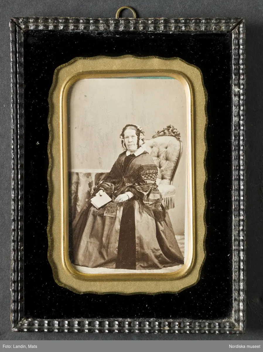 Porträtt i helfigur, fröken Maria Elisabeth Söderström (1782-1871), sittande i stoppad nyrokokofåtölj, mittbenat hår, iklädd liten mössa, mörk klänning med vid kjol och vit krage. Bord med duk på till vänster, håller en bok i höger hand. Eventuellt panotypi. Nordiska museet inv.nr 205018.