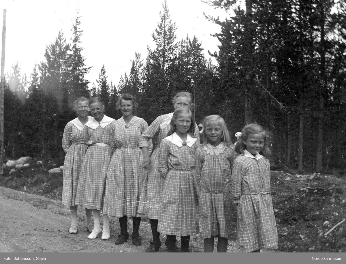 Gruppbild på skogsväg. Sju flickor i likadana rutiga klänningar står på rad.