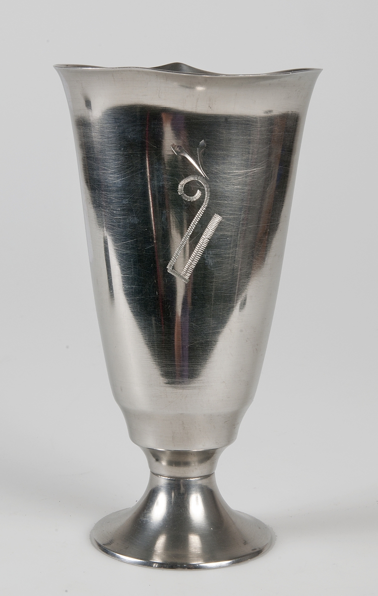 Metallbägare med fot. Smal kuppa, slät med ornament mitt på. Ingraverat: C. K. S. 1933.