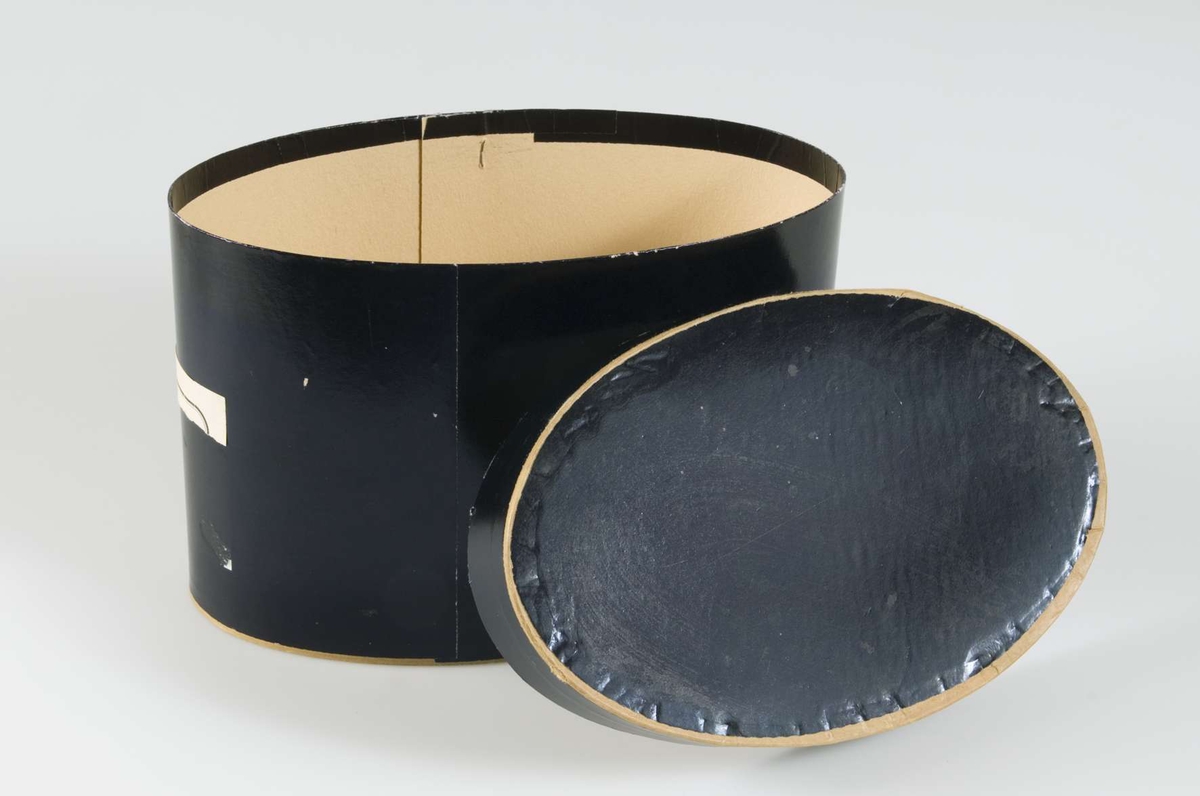 Oval ask med lock, av papp, klädd med svart papper.
Innehåller krimmermössa UM29562. 