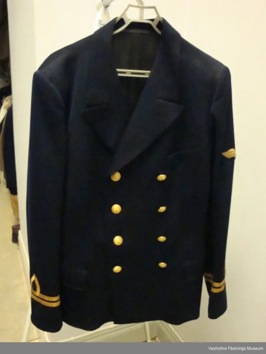 Uniform, kavaj och byxor i blått kläde m/FL, med löjtnants gradbeteckning. På höger ärm flygtecken m/15 (flygspanare)