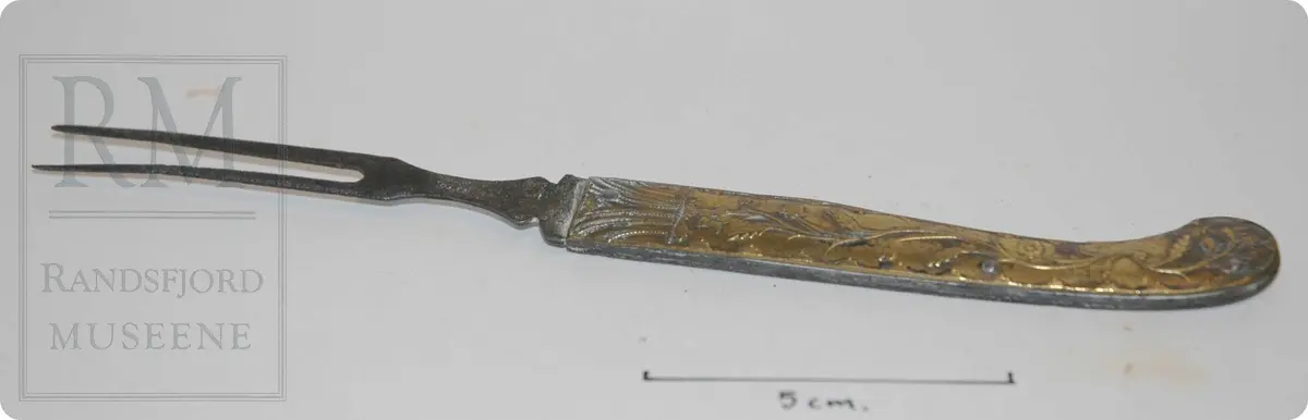 Sammenleggbare gaffel, som en foldekniv. Jerntenner, messingskaft, med trykket blomsterrankedekor. Gaffel har hengt sammen med kniv. Denne mangler. Se HF-00613 (komplett)