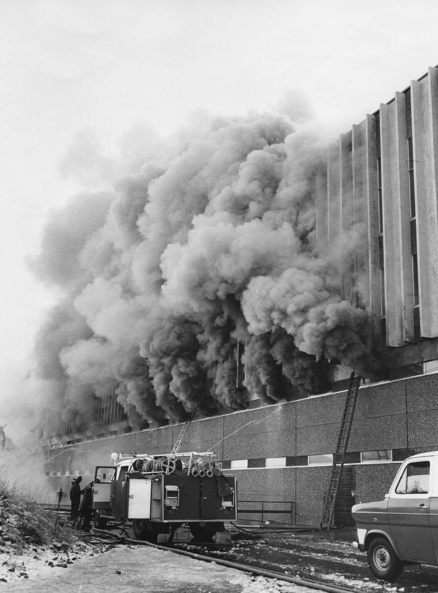 Brann ved sykkelfabrikken på Kvål, 16.11.1973: røyk kommer ut av bygningen, brannbil foran bygget