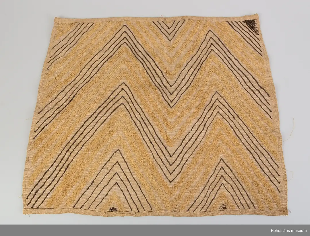 Text till webbutställning "Från när och fjärran" presenterad på Bohusläns museums hemsida år 2009 - 2013:
Vävnad av rafiabast med broderat mönster, delar av mönstren har flossakaraktär, 67 x 60 cm.
Mönstren, som täcker hela den vävda tuskaftsbottnen, består dels av linjer, dels av ytor med sammets- eller flossakaraktär. Typisk är att mönsterformerna plötsligt ändrar karaktär och symmetrin saboteras på ett befriande sätt. Baksidan är nästan enfärgad, broderiet syns endast i svag relief. 
Basten kommer från rafiapalmens långa blad, Rafhia farinifera. Bladen kan bli upp till 20 meter långa. I Sverige träffar vi på materialet under benämningen trädgårdsmästarbast. 
Textilier av detta slag utfördes i Kasai-området i södra delen av Belgiska Kongo, nuvarande Demokratiska republiken Kongo. Där fanns från 1600-talet Kuba-riket med Shoowa-stammen, som var känd för sin mycket speciella och högt skattade broderiteknik. Det var männen som vävde, medan kvinnorna broderade de geometriska mönstren i dessa prestigetextilier, ibland använda som betalningsmedel.
Gåva av Sjökaptenen och Riddaren Elmer Göransson år 1961.

Uppgifter om givaren
Under sin 30-åriga vistelse i Belgiska Kongo skötte Elmer Göransson (1872-1964) under många år transporter på landets floder. När han år 1932 avslutade sin tjänstgöring var han både skeppsfartygsinspektör för hela den belgiska flottan i övre Kongo, t. f. chef för hydrografiska tjänsten samt hamnkapten i Leopoldville.
Han var samlare av bl.a. vapen, textilier och träkärl. I omgångar från 1919 skänkte Göransson afrikanska föremål till Uddevalla museum. Han kom att bli en av museiföreningens hedersledamöter.
Vid sin bortgång 1964 var han bosatt i Kungsbacka.

Artikel i Bohusläningen från måndagen den 1 november 1937.
"Vittberesta bohusläningar berätta. Upplevelser under 30-årig vistelse i Belgiska Kongo. Kapten Elmer Göransson. Folk och sedvänjor under ekvatorns glödande sol."
_______________________________________________________________________

Kontinent: Afrika
Information på lappkatalogkort:
Matta, 67 x 60 cm. Geometriskt mönster i gult och brunt på blekskär botten. Av rafia. Flossaliknande. Teknik som 61.03.076. Congo, Afrika

Enligt litteraturen är vävnader av detta slaget utförda av stammar som levde i området Kasai i södra delen av forna Belgiska Kongo, senare Zaire. Man pratar om det på 1600-talet bildade Kuba-riket. Ett folk inom Kuba, Shoowa-stammen, var kända för sin mycket speciella och högt skattade broderiteknik.
Det var männen i Shoowa-stammen som vävde, medan kvinnorna broderade de geometriska mönstren.

Sicksackformat broderat mönster i gult och brunt mot ljusare gulvit tuskaftsbotten. Flossakaraktär på delar av broderiet.
Stadkanterna på vävnaden är vikta och fållade mot framsidan med förstygn; i längdriktningen är fållen vänd mot baksidan och sydd med förstygn.

Litteratur: Artikel i Hemslöjden nr 4/1990, "Den saboterade symmetrin", Olle Nessle, s. 14-15.

Uppgifter om E. Göransson i Knut Adrian Anderssons katalog samt historik om samlingen: Se UM61.03.001

In English: Cut-pile raphia cloth, the so-called Kasai velvets. Bakuba, Zaire.