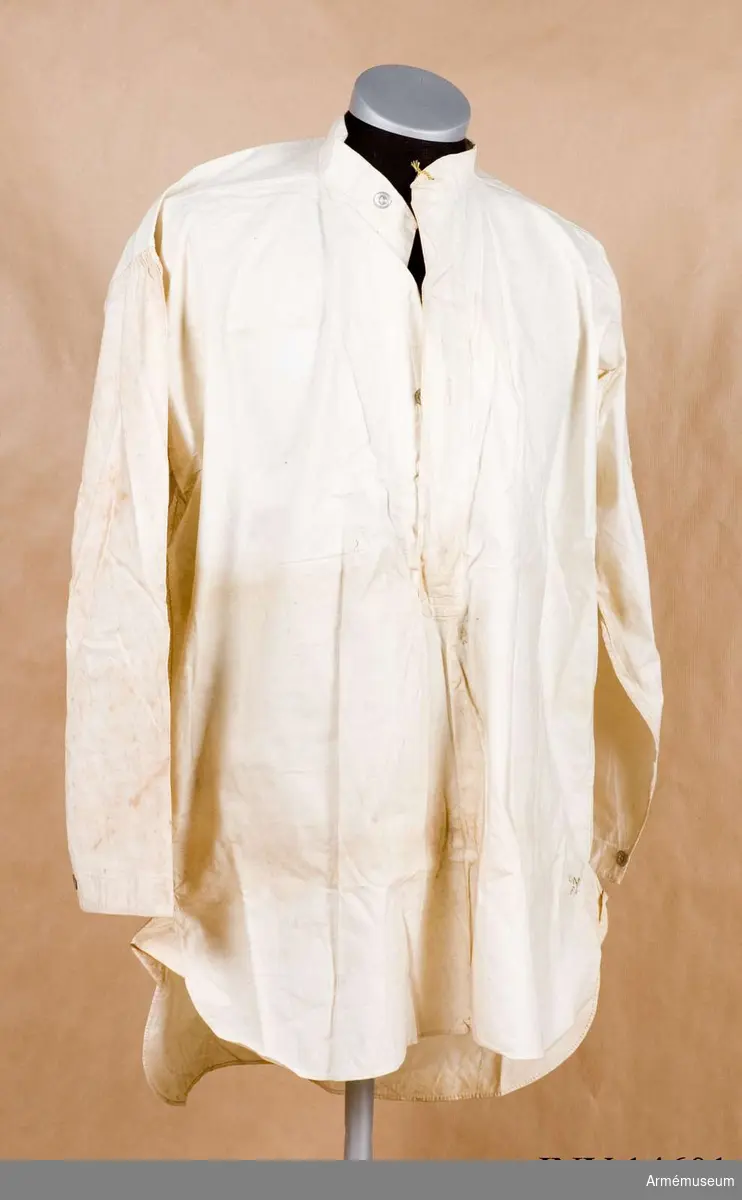 Skjorta av vitt bomullstyg med fast upprättstående krage med långa ärmar. Kragen och bröstöppningen knäppes med två metallknappar. På varje ärmuppslags sprund en knapp av samma sort. PÅ skjortans framsida stämpel: "W" upprättstående pil "D", "13" under pilen.