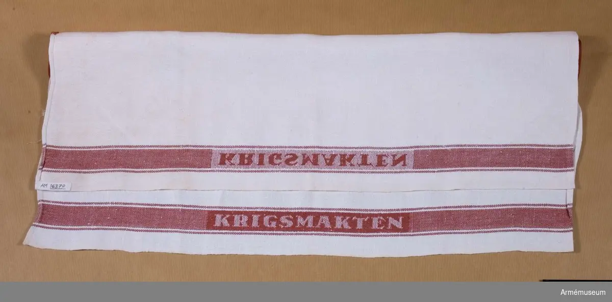 Samhörande nr är 16376-16379.Handduk.Handduk i halvlinne vävd i kypertteknik. Vit med röd bård runt om. På båda långsidorna i den röda bården är ordet KRIGSMAKTEN invävt.Handduken funnen i museet vid inventering av förråden 1987-07-28. Den är tillverkad före år 1975 då namnet Krigsmakten ändrades till Försvarsmakten.