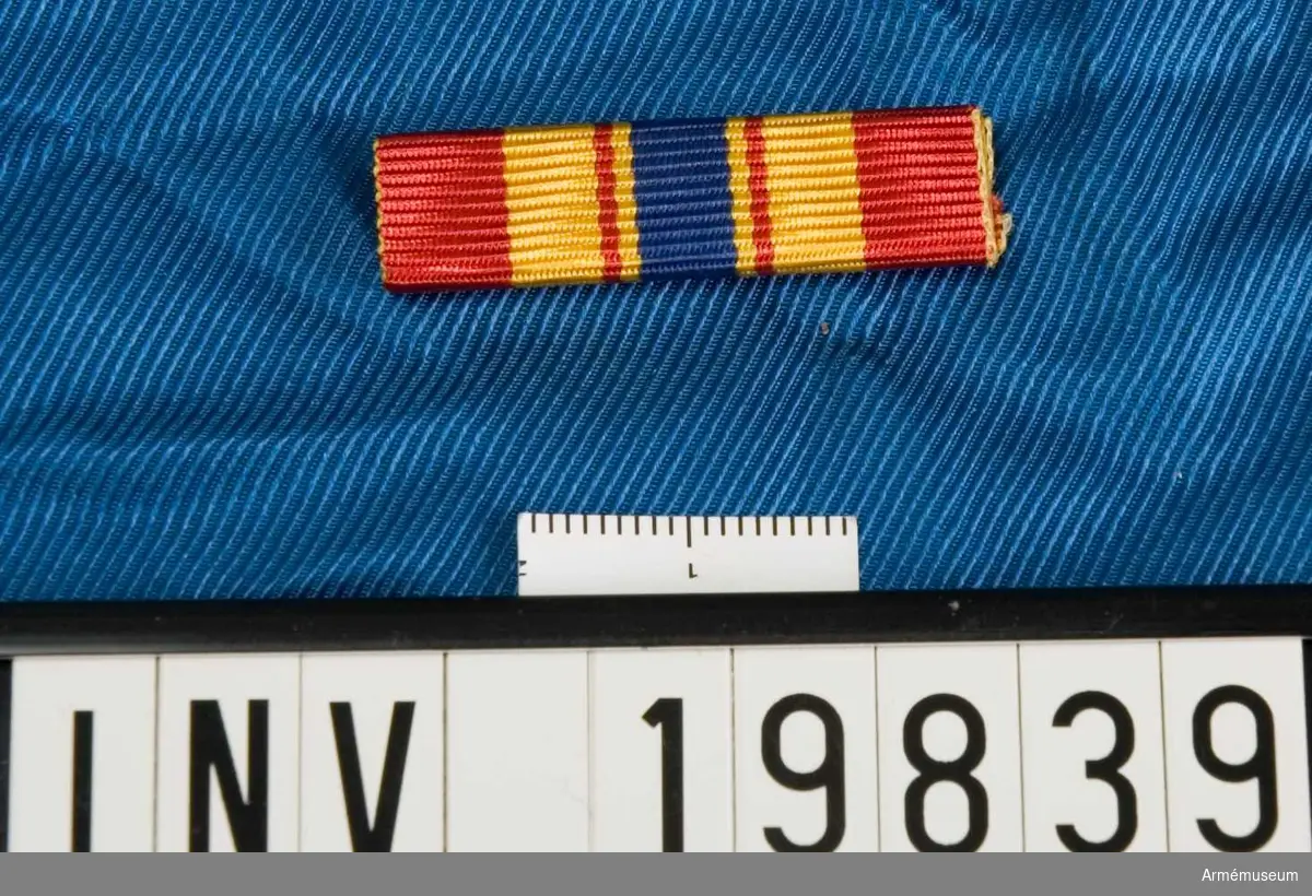 Gult band med breda röda kanter, ett blått band i mitten med ett rött streck på vardera sidan. Släpspännet förvaras i en ask tillsammans med en medalj och en miniatyrmedalj.