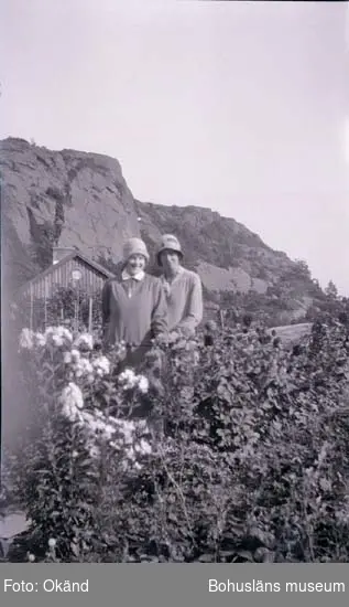 Två kvinnor klädda i klänning och hatt står bakom en blommande rabatt. 
I bakgrunden ser man ett boningshus och längre bort ser man ett berg.