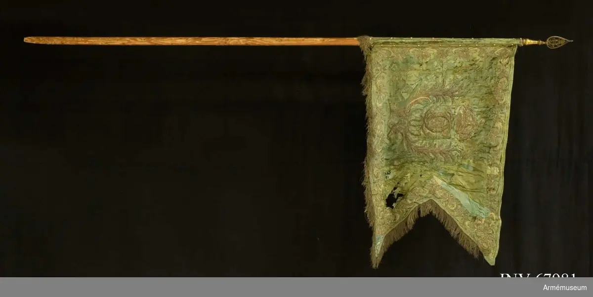 Duk: Tillverkad av dubbel grön sidendamast, tvåtungad, sydd av två horisontella våder. Kantad med frans i silver (guld). Fäst vid stången med förgyllda spikar på guldband. 

Dekor: Broderi på båda sidor i silver (guld) lika på båda sidor, Karl XII:s namnchiffer, dubbelt C, därinom XII, under sluten krona, innefattande av, två, nedtill korsade lyrformigt böjda palmkvistar, nedtill hopbundna med rosett. Längs kanten bård med rika ornament.

Stång av målad grön furu. Kanellerad. Holk av förgylld mässing. Spets med Karl XII:s krönta namnchiffer.

Anteckning från katalogkort:
I spetsen på inv.nr 153 var inlagd en bit av en dagorder för Bohusläns infanteriregemente från 1808.