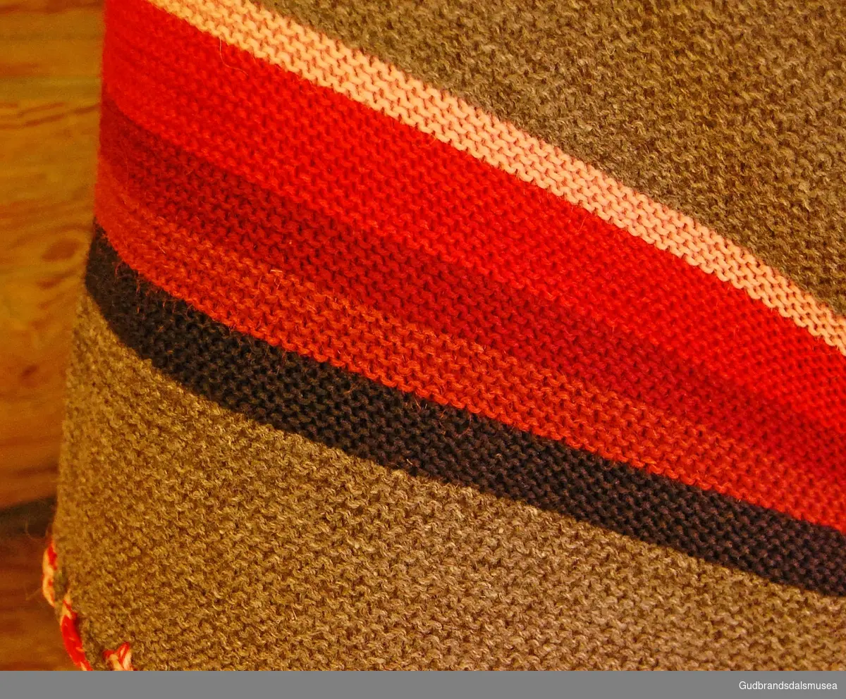 Bondetørkle i strikket ull, med striper i ulike farger, knyttet bånd i rødt og hvitt rundt halve tørklet, heklet snor. To nåler i metall med lenker til å sette sammen/feste tørklet ved bruk. 