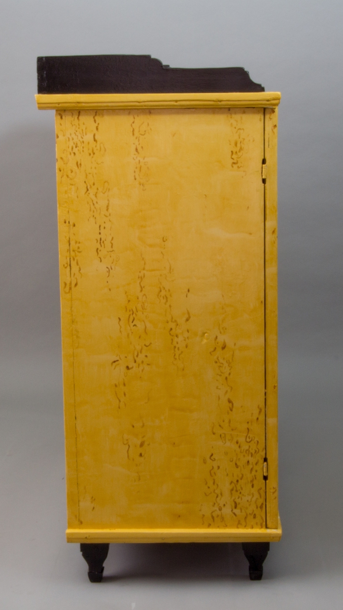 Skänk tillverkad av furu. Upptill runt skivan löper en svartmålad kant. Möbeln har raka sidor och en framsida med två dörrar. Dörrarnas speglar är dekorerade med kannelerade romber. Innanför dörrarna döljer sig en hyllinredning och en större låda upptill. Skänken vilar på kannelerade fötter fram. Möbeln är målad med en björkimitation flammig i gula toner, vissa detaljer är svärtade.