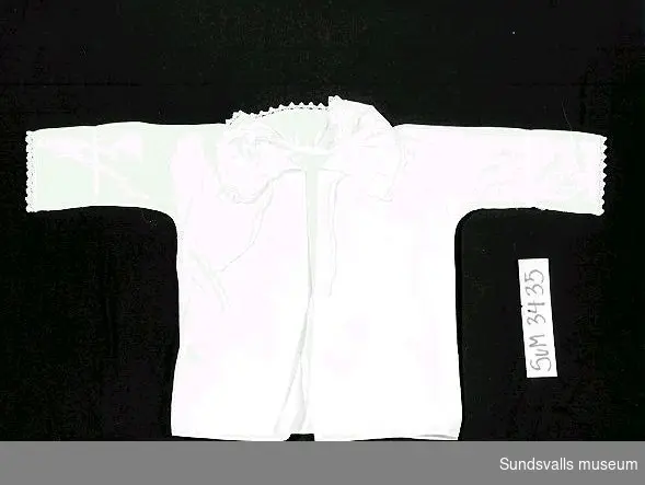 Babyskjorta i vit bomull med liten spets kring krage och ärmar. Knytes.