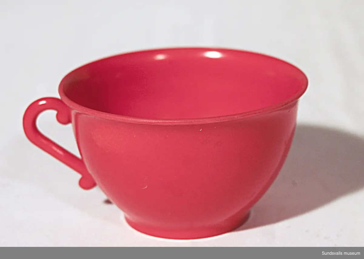 SuM 5240:1-4 del av leksaksservis i röd plast. SuM 5240:1 kaffekanna med lock. Höjd 10,5 cm. SuM 5240:2-3 kaffekopp och fat. SuM 5240:4 kaffefat. Diam. 9,8 cm.