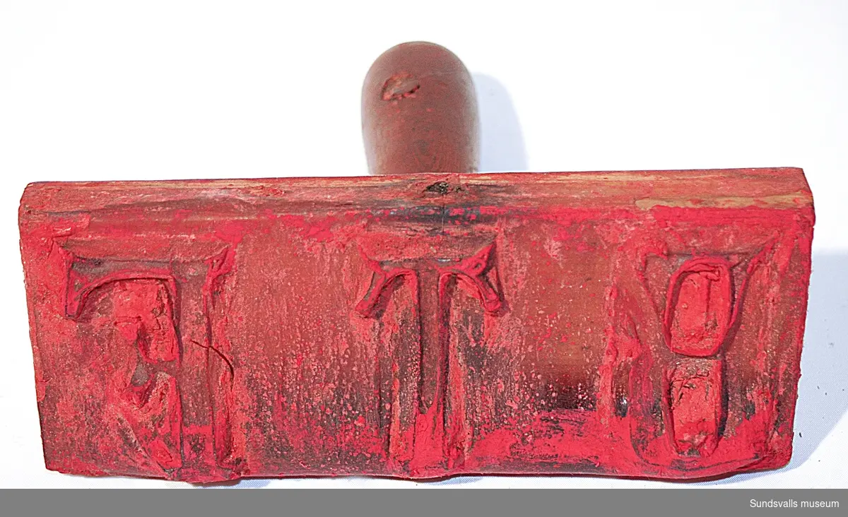 Stämpel i gummi med handtag i trä. Stämpeln visar bokstäverna BT. Röda färgrester finns kvar på stämpeln.