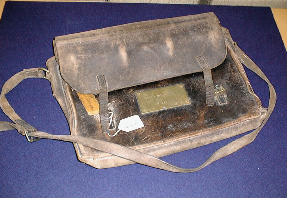 Lösväska i läder med klaff och två spännen. På framsidan en mässningsbricka med inskription i skrivstil. Väskan är försedd med låsten, hänglås och bärrem.