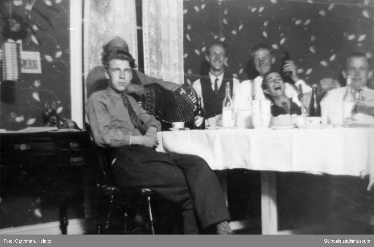 Fest i Fjärås Bräcka, 1930-tal. Sex män sitter samlade runt ett flaskfyllt bord. En man spelar dragspel och personen längst fram heter Bror Eriksson.