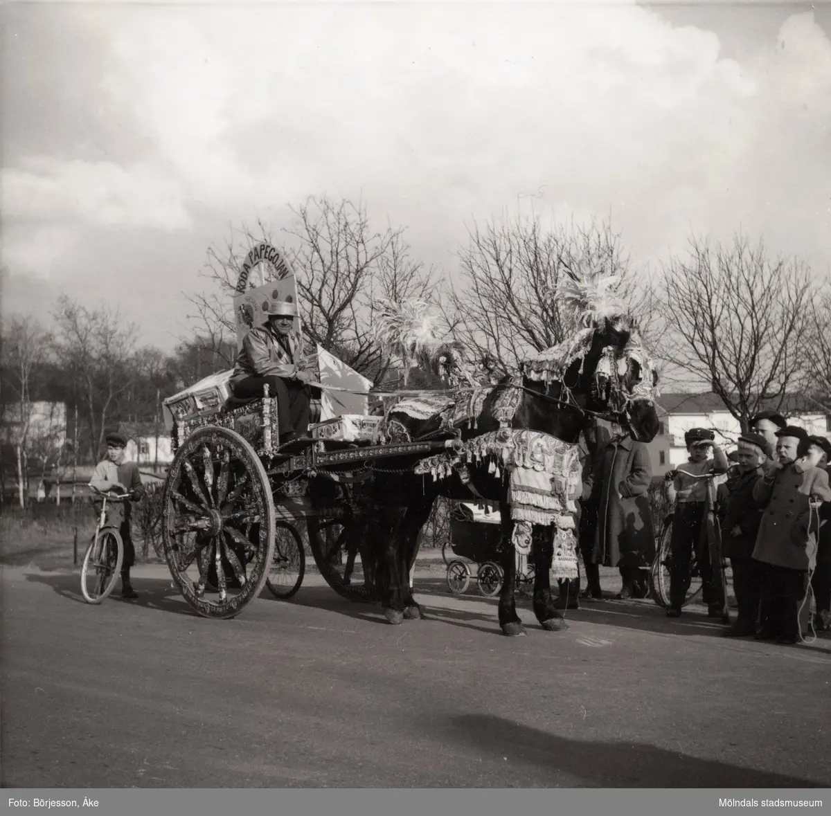 Motiv från Solängen. Häst med vagn och kusk bär dekorationer. Möjligtvis reklam av något slag? Skylt med texten "Röda papegojan" på vagnen. Barn på cyklar står runt omkring och tittar på.
Möjligen Frölundagatan på 1950-60-talet.
