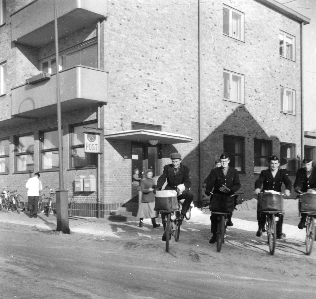 Cyklande lokalbrevbärare i Ljungby på väg ut med post i april 1955. I bakgrunden postkontoret.