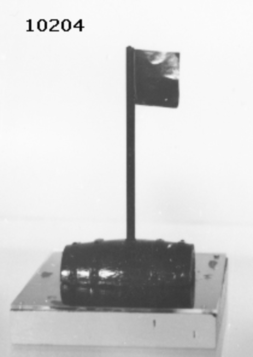 Modell av "Vrakboj". Tunnan är försedd med fyra stycken band samt en stång med en flagga av plast. Grönmålad. Bojen är monterad på en träplatta, botten och sidorna är ljusgrå, översidan blå, täckt av ett plexiglas.
