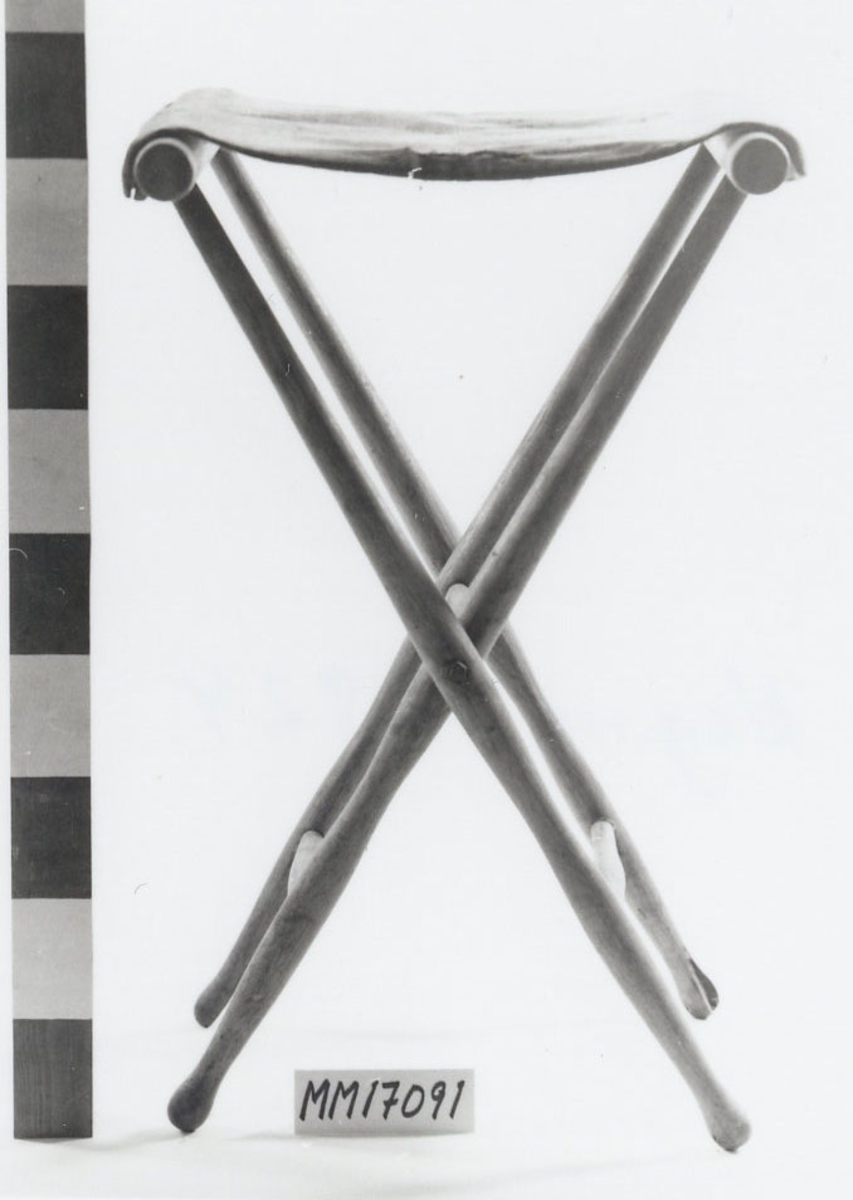 Fällstol av ek? Fyra runda, svarvade ben, fram- och bakben hålls samman av tvärslåar nederst samt överst där tvärslån även fungerar som sida till sitsen. Vänster och höger benpar är korsvis fästa i varandra med tvärslå, och kan fällas ihop. Sitsen består av rekangulärt tygstycke av grön sammet (?) förstärkt på baksidan med segelduk. På kortsidan försedd med läderband som med hjälp av knapphål knäppes fast på fem mässingsknappar som är indrivna i tvärslåns utsida.