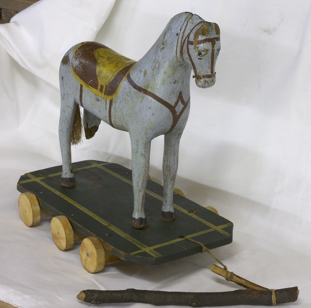 En lekehest i tre. Mangler ett bein. Står på en platting med hjul. Hesten er malt lyseblå og har påmalt sadel, hodelag og brystreim. Har med en liten"tømmerstokk" til å trekke den med.