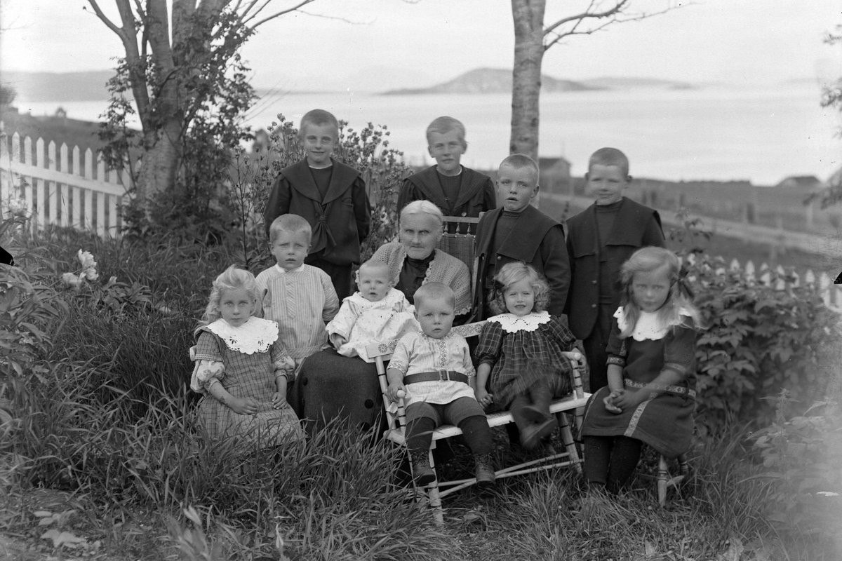 Eldre kvinne og ti barn, fotografert i hagen på Røkenes gård. Kjeøya i bakgrunnen.