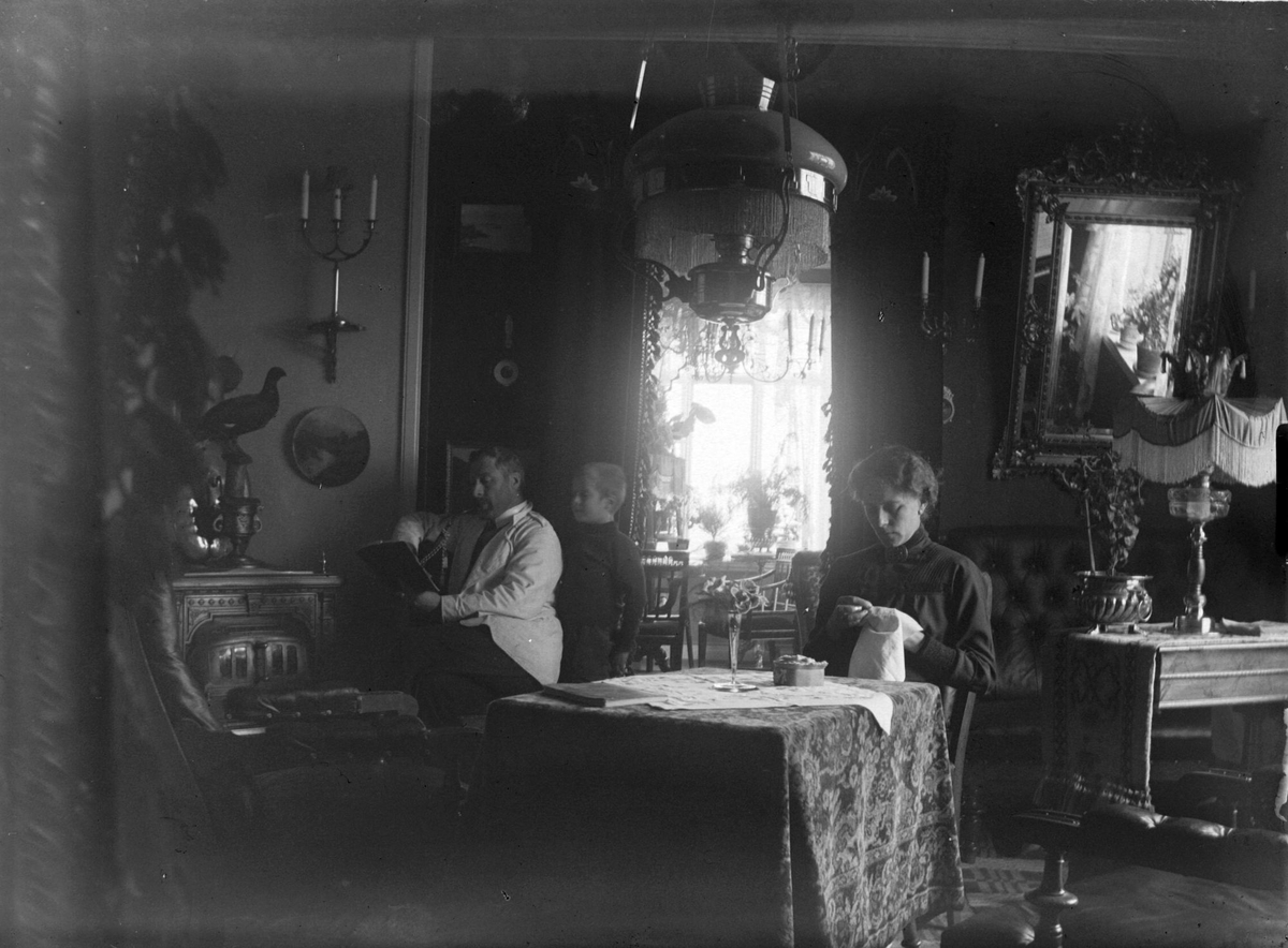 Familien til lege Christian F. Rossow fotografert i stua. Til venstre leser doktoren en bok, mens sønnen Eivind ser over skulderen hans. Kona Dagny sitter til høyre og broderer.