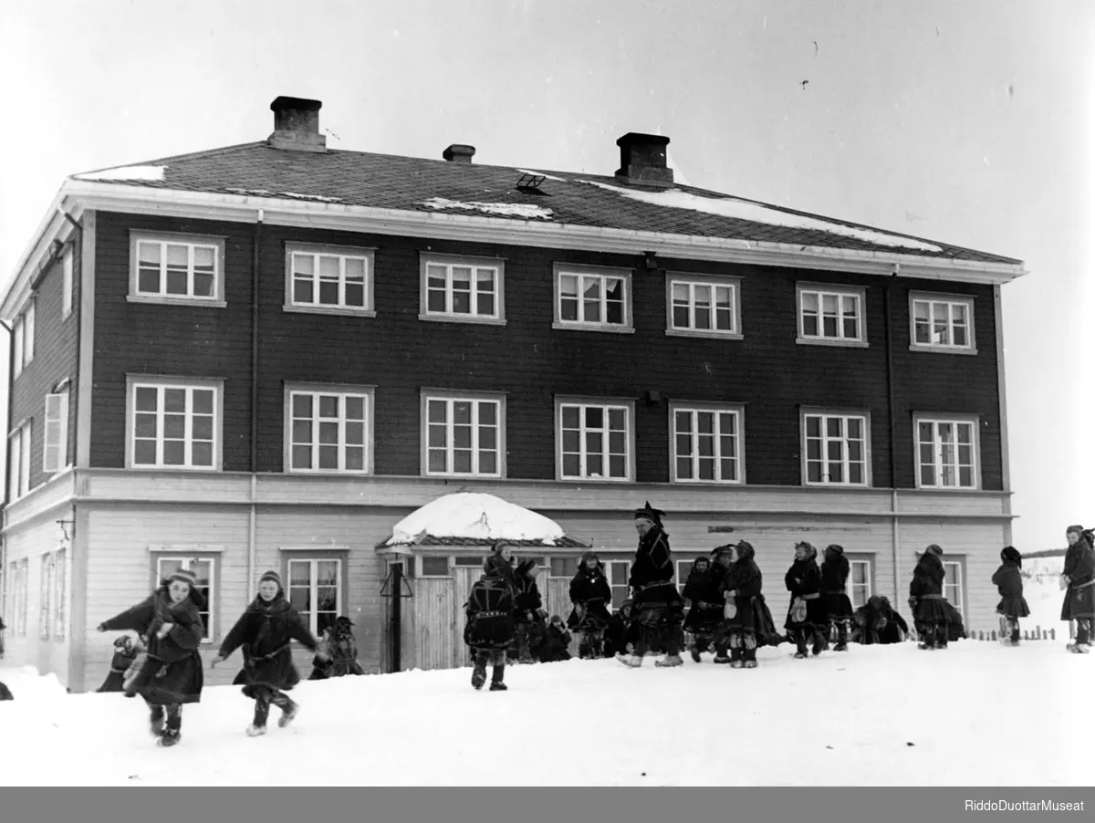 Gávtehas mánát stohket skuvlašiljus 1935.
Koftkledde barn leker på skoleplassen 1935.