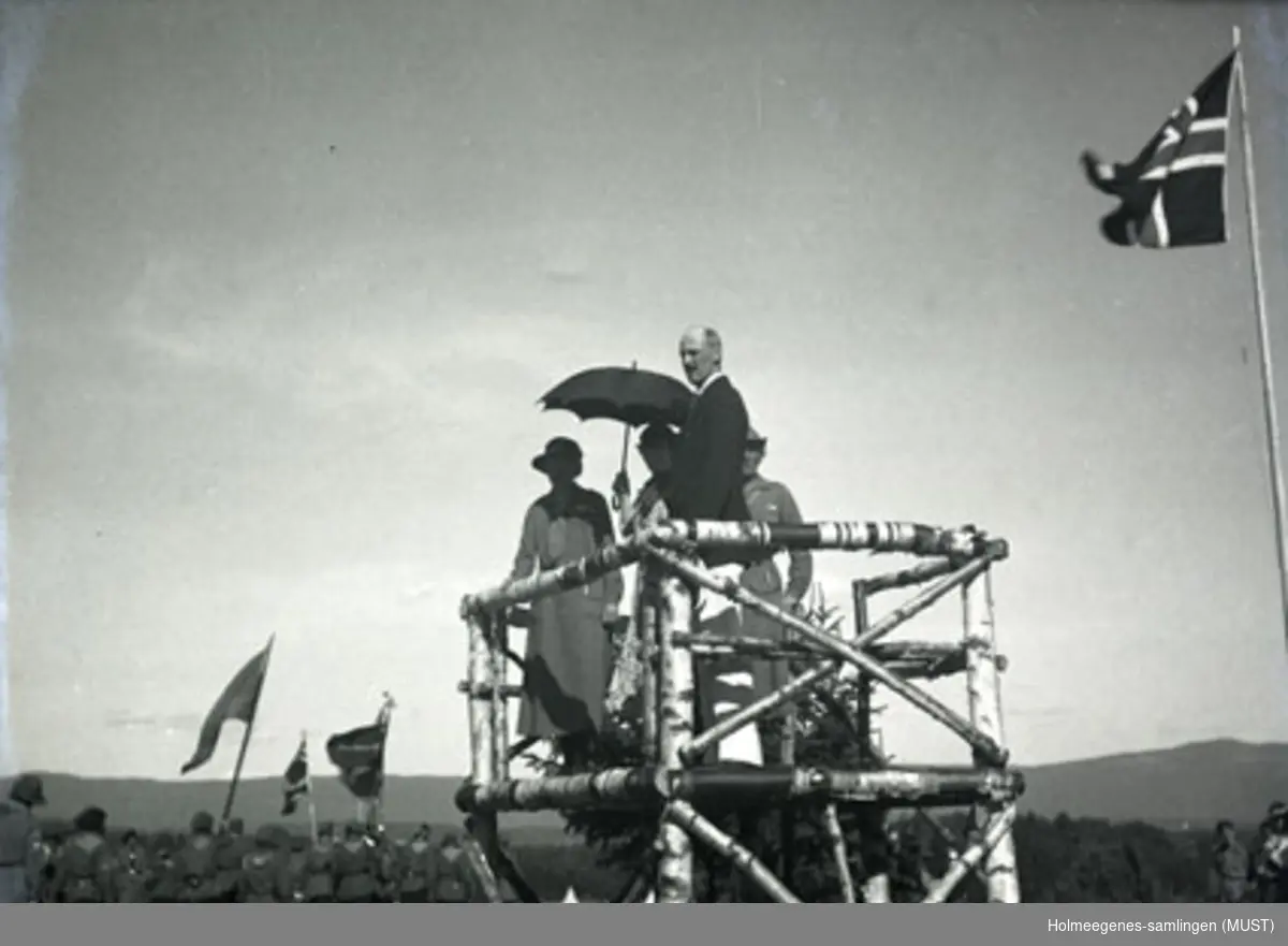 Kong Haakon VII, dronning Maud og en kvinne og mann på en talerstol (podium) bygd av bjørkestammer. Landsleir på Ringerike 1935. Den ukjente kvinnen er antatt å være leirens leder, Josefine (Joss) Barman.