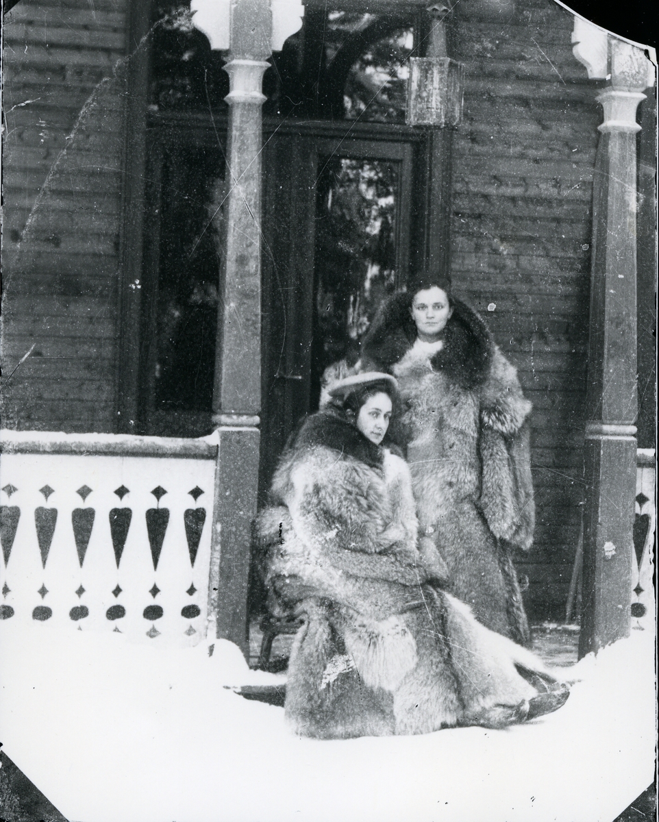 To kvinner kledd i store pelskåper, i inngangsparti til sveitserstilsbygning