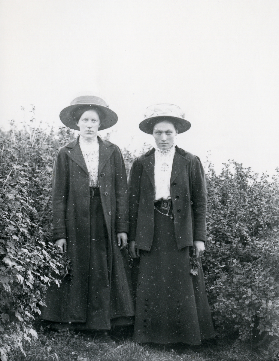 To kvinner med hatt, i helfigur, stående foran bærbusker