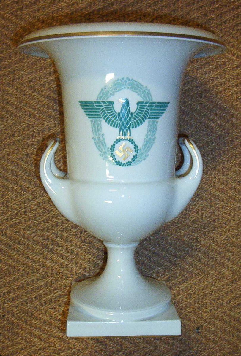 Pokalformet urne i hvitt porselen. Dekor i gull og grønt. Urnen bærer det tyske ordenspolitiets merke, en omkranset tysk ørn over hakekors.