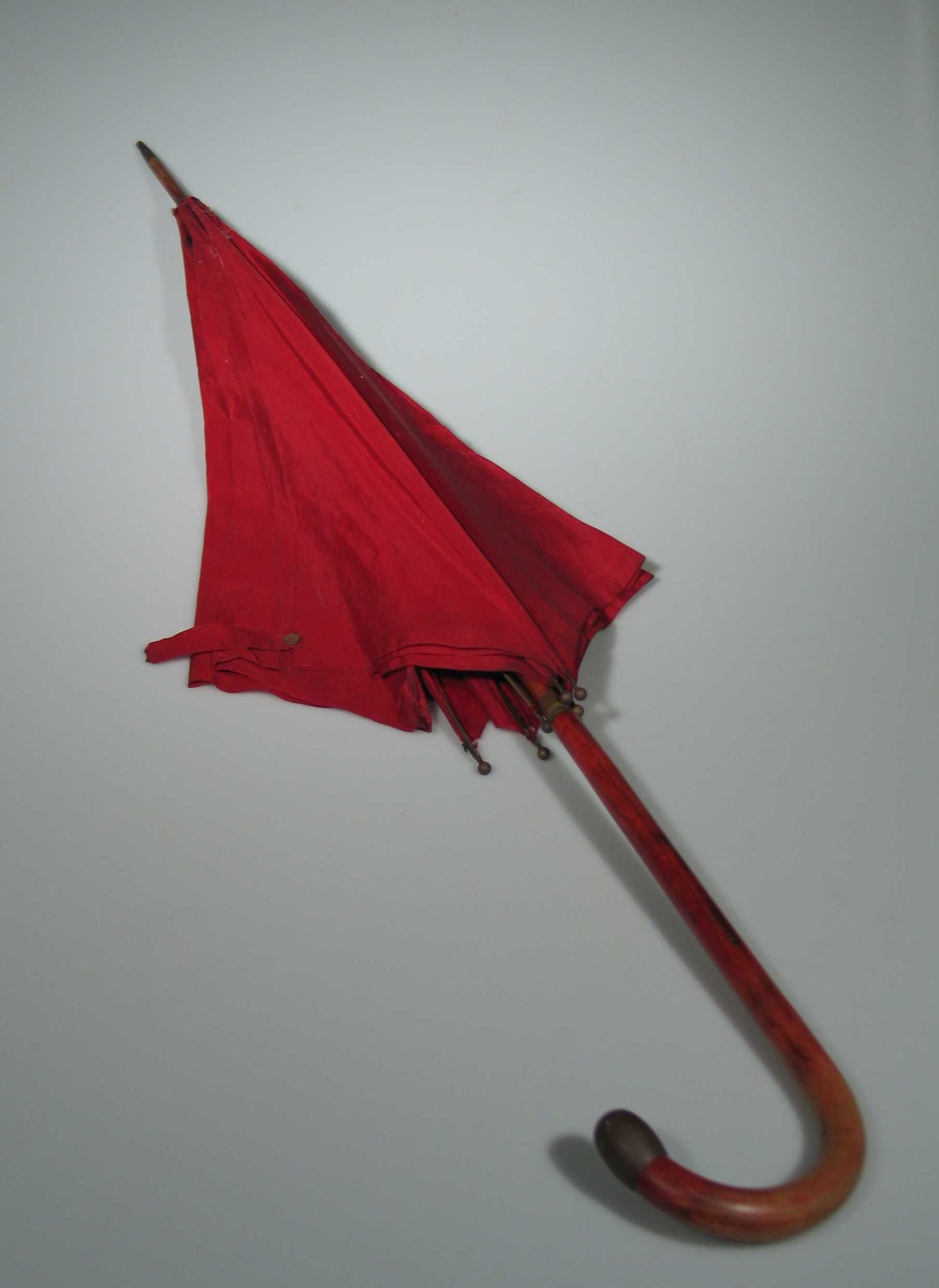 Paraplystang av tre med buet håndtak. Messingholk øverst og nederst og messinkule, ca 6 mm i diam på toppen av hver spile. Messing i låsmekanisme. Rød to-skaft silke i skjerm.