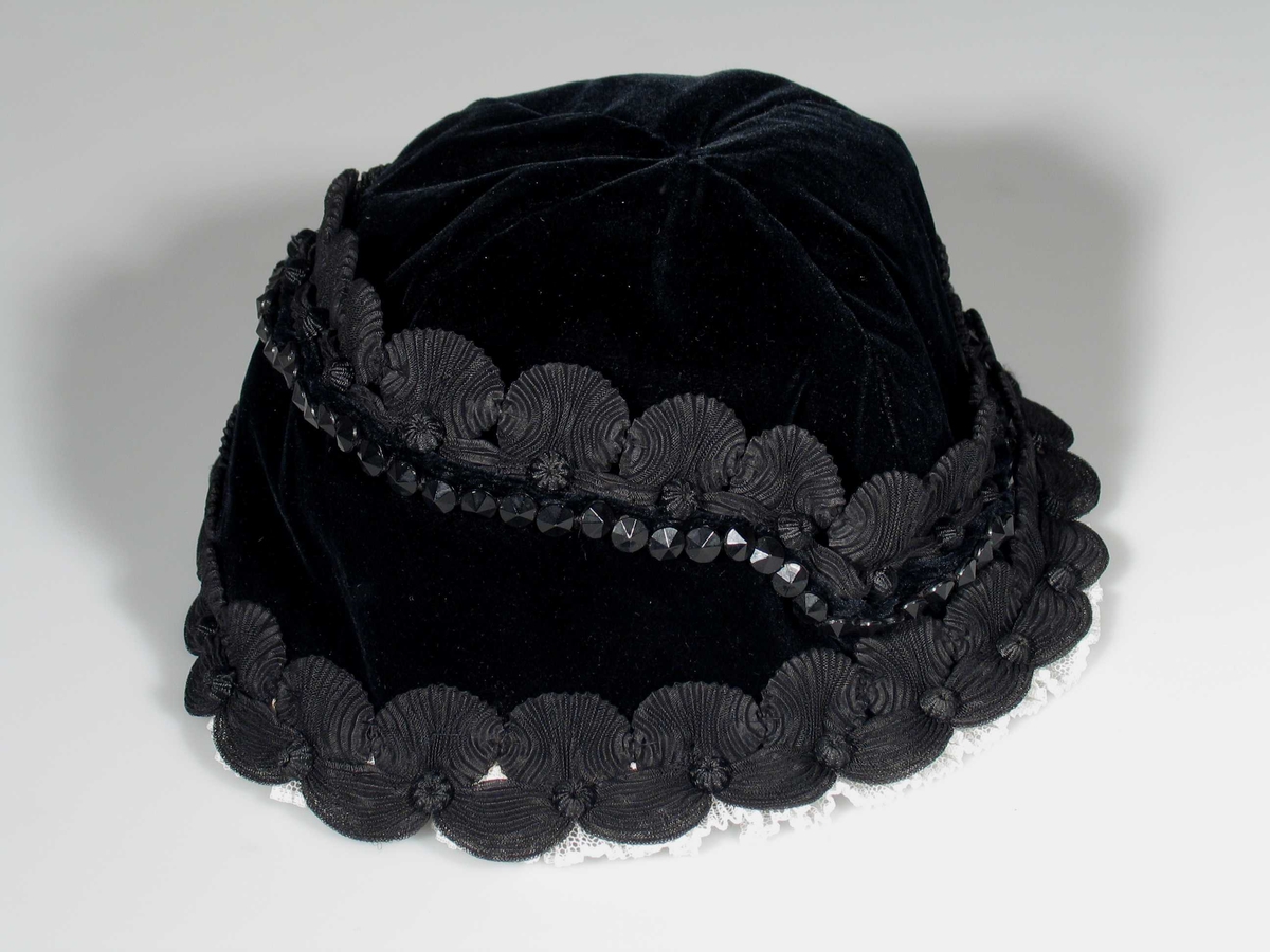 Svart hatt i fløyel kantet med plisserte bånd og steiner. Foret med svart silke.