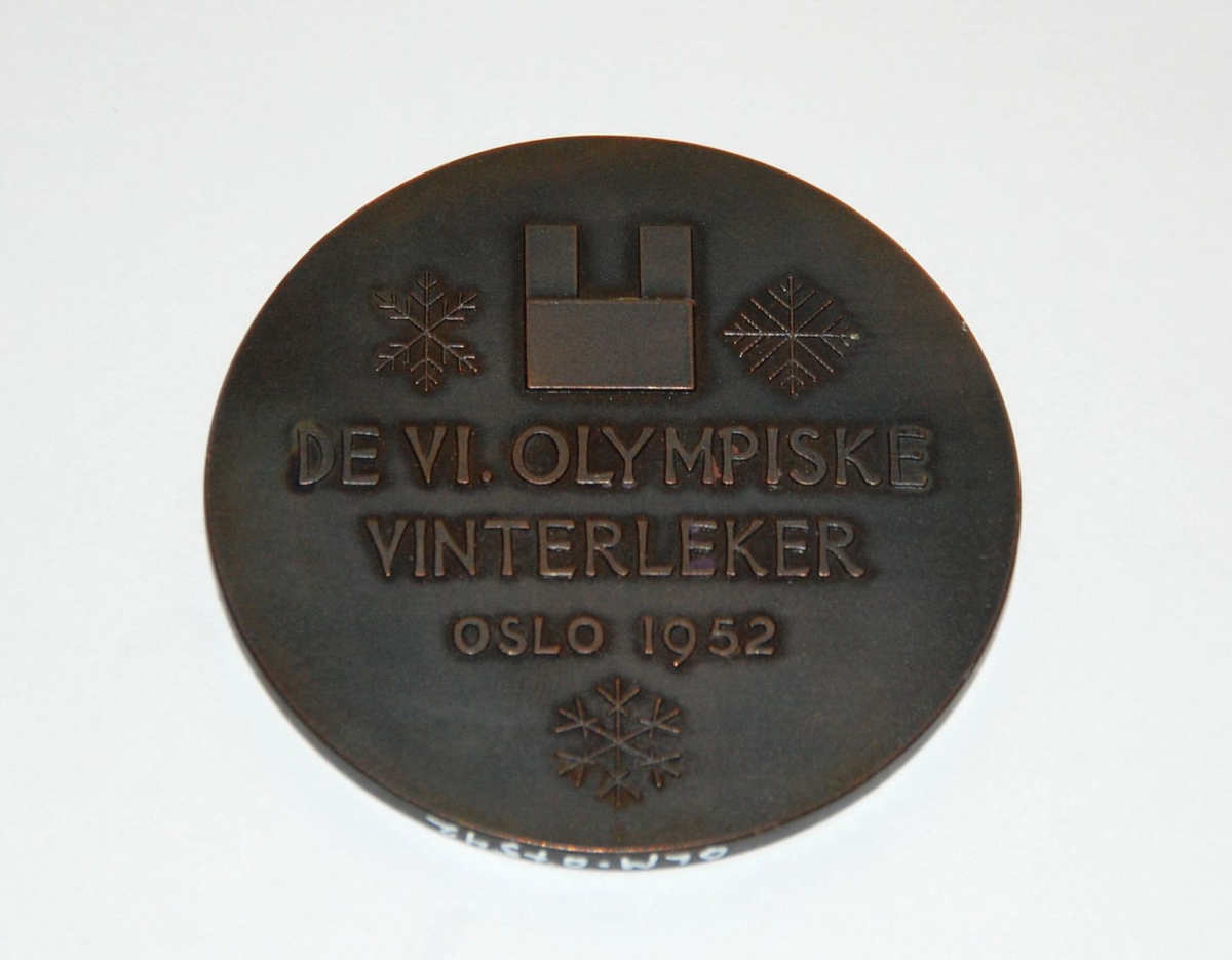 Bronsefarger medalje. På den ene siden er det motiv av rådhuset i Oslo og snøkrystaller. På den andre siden er det motiv av en fakkel og de olympiske ringene.