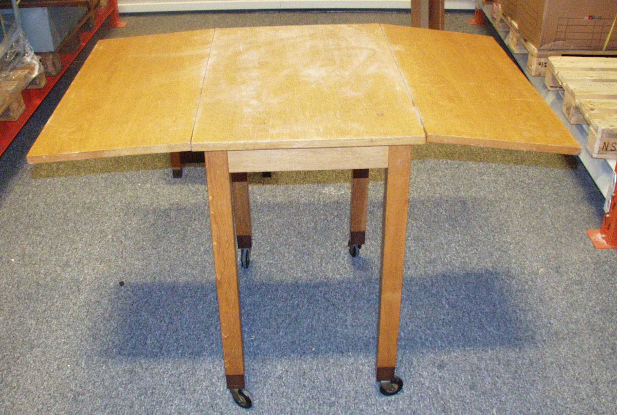 Bord i tre. Det er med klaffer som kan slåes opp, slik at bordet blir større. Bordet har fire ben med hjul på. 