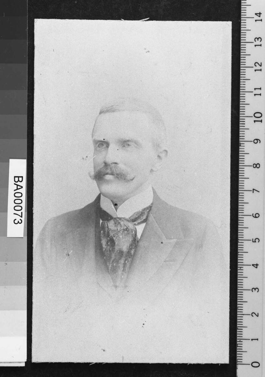 Portrettfotografi av en mann med snurrebart og stort silkesjerf rundt den hvite opprettstående skjortekrave og mørk dress med vest.