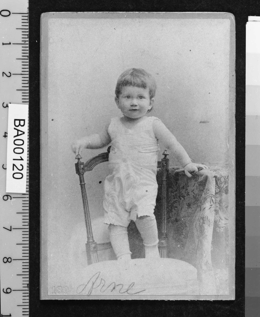 Barneportrett, liten gutt i hvitt undertøy stående på stol, venstre hånd støtter på bordkant. Han ser direkte på betrakteren.