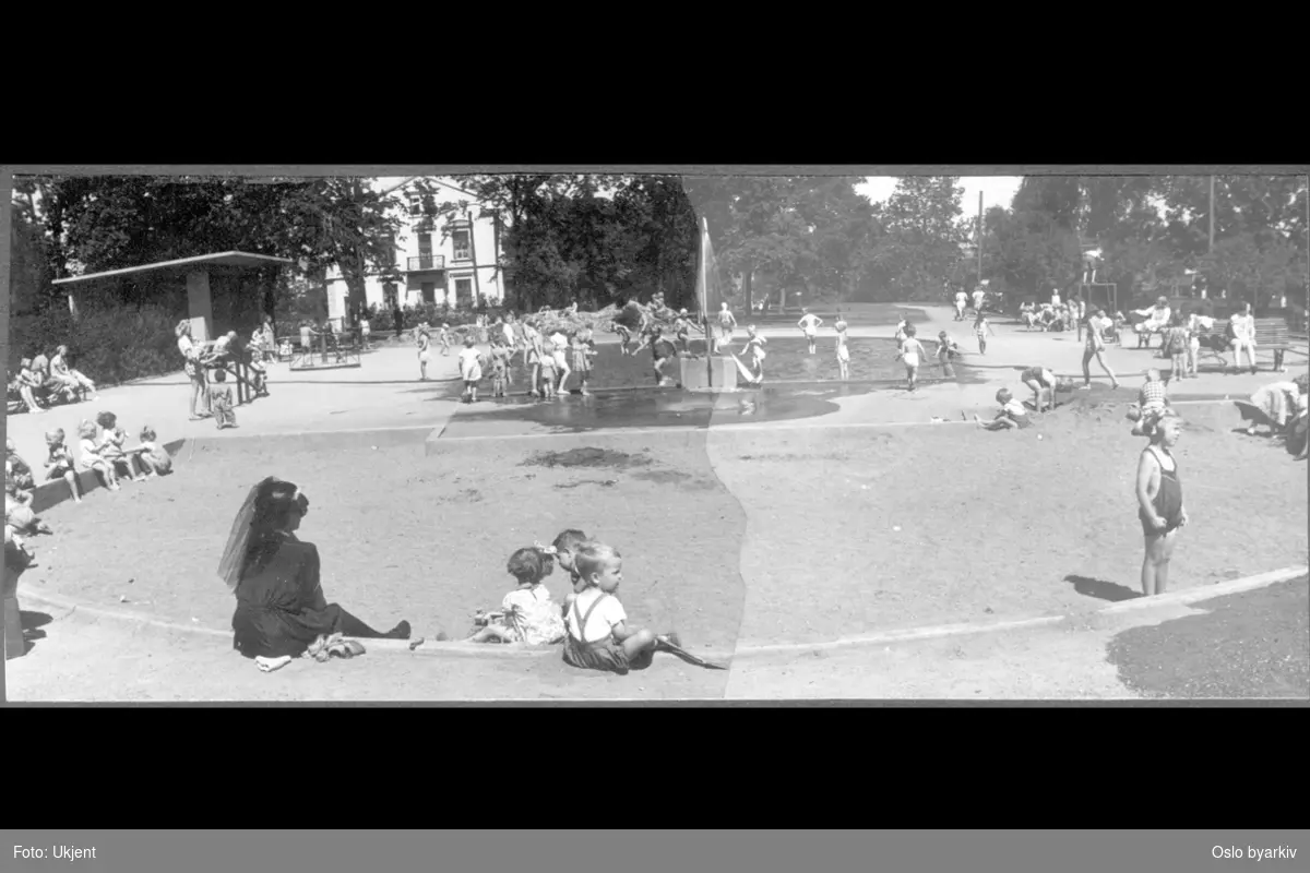 Lekende barn og mødre på øverste lekeplassen i parken. Vassebasseng / Plaskedam med dusj. Sandkasse. Leskuret til venstre.