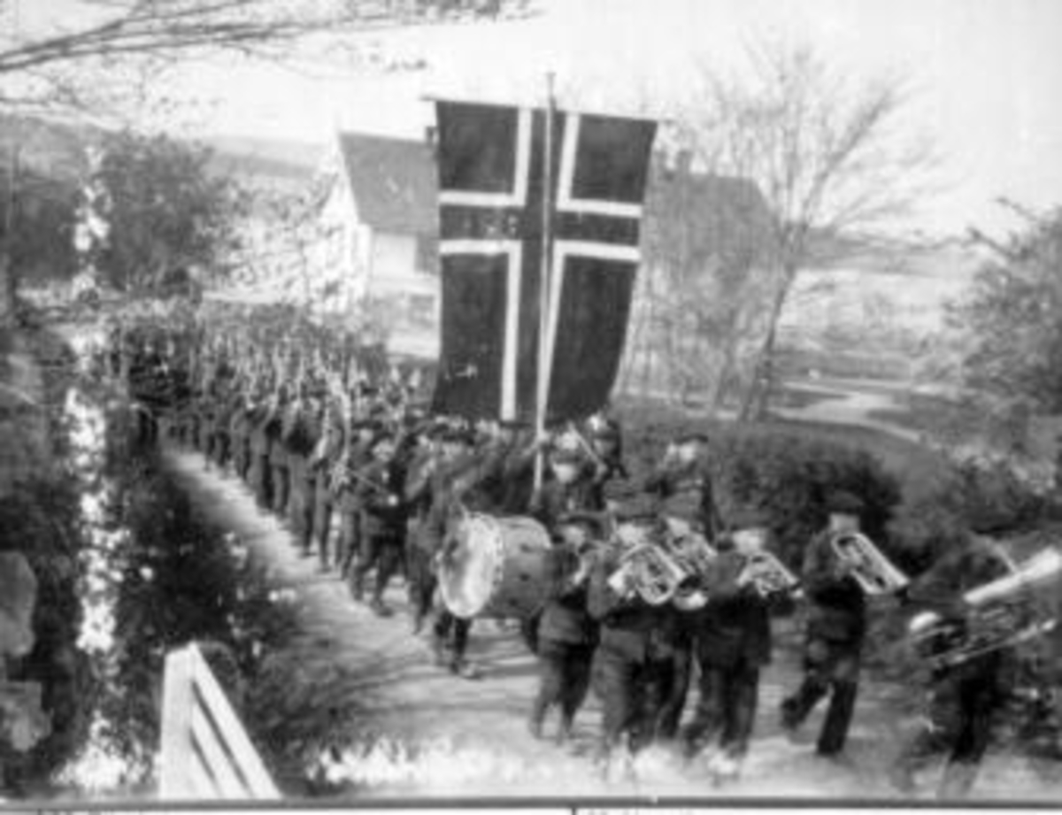 17. mai-toget marsjerer over tunet på Toftes Gave, Nedre Sund, Helgøya med orkester i front. Mange mennesker og flagg.