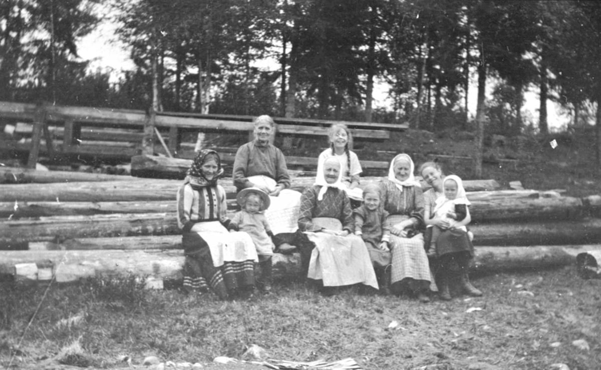 Kvinner og barn sitter på tømmerstokker på Gjerlaugbakken gård, Furnes.