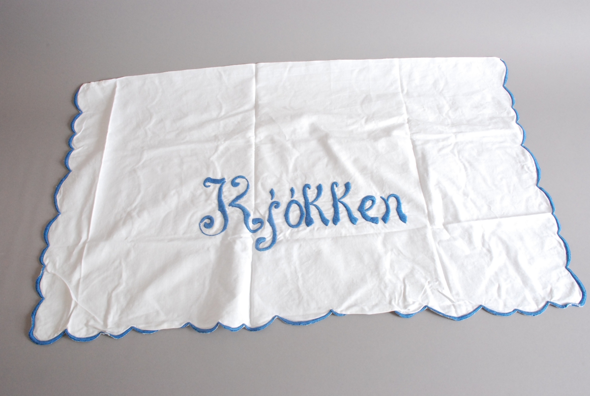 Hvitt pyntehåndkle med blå broderte render tre sider. Tekst:  "Kjøkken" brodert med blå tråd.