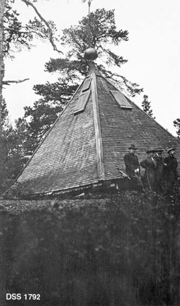 Takkonstruksjonen til utsiktstårnet på Fjellheim i Meldal, nedblåst etter kraftig uvær.  Takkonstruksjonen har pyramidal form, og er tekket med trestikker.  Øverst på takflatene er det treluker, og helt i toppunktet er det festet en metallkuppel.  Takkonstruksjonen ligger på bakken.  Fire herrer sitter på takets ytterkant.  Fjellheim var sommerstedet til industrilederen og forretningsmannen Ole Christian Marentius Thullin Thams (1837-1907). 